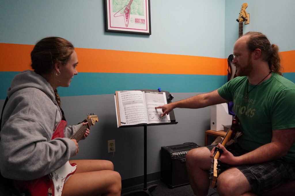 A student learning to play on electric guitar in front of a teacher.