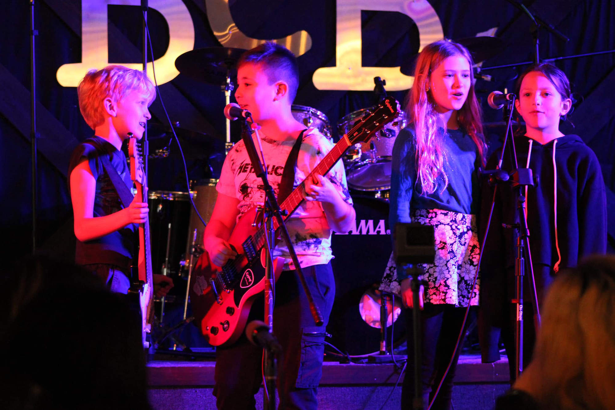 Four children holding instruments performing on stage and singing into two microphones.