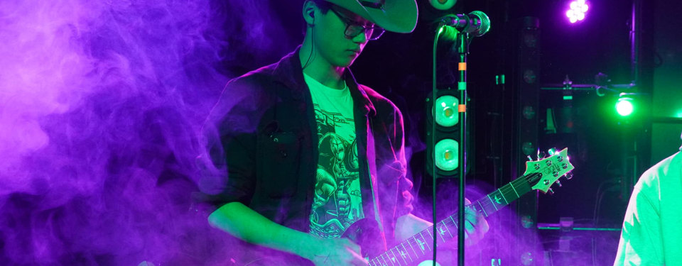 Student in a cowboy hat playing guitar on a stage filled with colored fog.