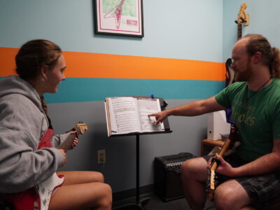 Guitar A student learning to play on electric guitar in front of a teacher.