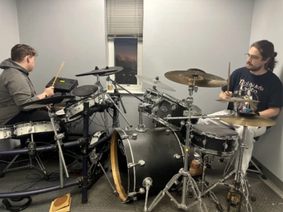 Drums Photo of a student practicing on an electronic drum set while their teacher plays on an acoustic drum set.